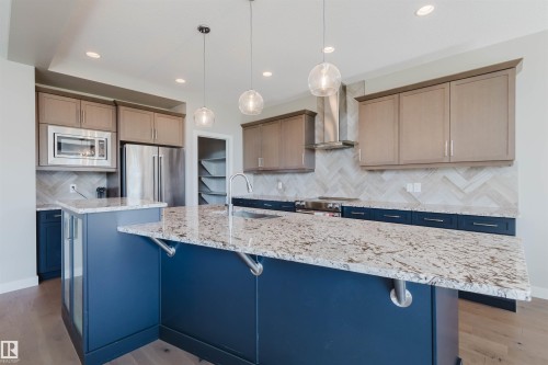 Kitchen featuring a large island with a speckled stone countertop, two-tone cabinetry, stainless steel appliances, a herringbone tile backsplash, and wood-finish flooring - 4125 Kennedy Green Sw, Edmonton, AB - Indoor Photo Showing Kitchen With Upgraded Kitchen