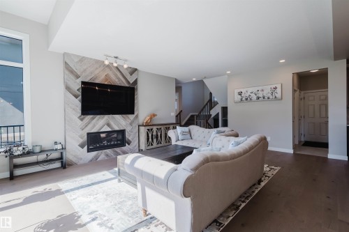 Living area featuring a fireplace with a herringbone tile surround, wall-mounted television, large window, and wood-finish flooring - 4125 Kennedy Green Sw, Edmonton, AB - Indoor Photo Showing Living Room With Fireplace