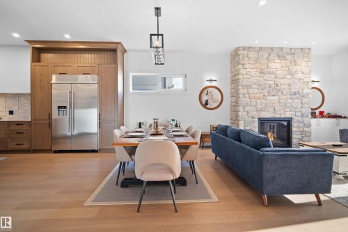 Open-concept living area with wood-finish flooring, a stone fireplace, recessed lighting, a modern linear chandelier, and integrated kitchen cabinetry featuring a stainless steel refrigerator - 12304 62 Ave Nw, Edmonton, AB - Indoor With Fireplace