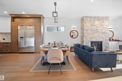 Open-concept living area with wood-finish flooring, a stone fireplace, recessed lighting, a modern linear chandelier, and integrated kitchen cabinetry featuring a stainless steel refrigerator - 
