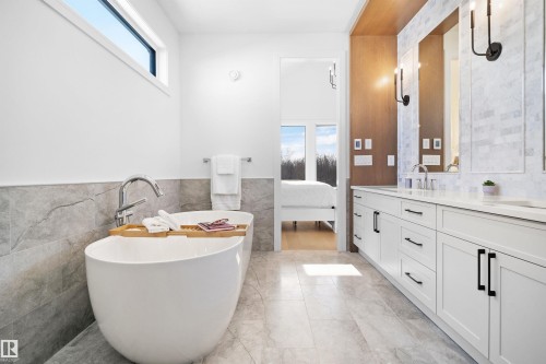 Modern bathroom featuring a freestanding soaking tub with floor-mounted chrome faucet, a dual vanity with white cabinetry and black hardware, and an elongated transom window - 12304 62 Ave Nw, Edmonton, AB - Indoor Photo Showing Bathroom
