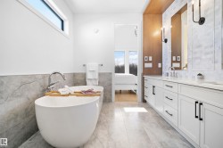 Modern bathroom featuring a freestanding soaking tub with floor-mounted chrome faucet, a dual vanity with white cabinetry and black hardware, and an elongated transom window - 