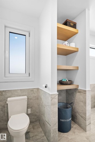 Bathroom featuring marble-look wall and floor tiling, a white toilet, a white framed window, and built-in wood-finish shelving - 12304 62 Ave Nw, Edmonton, AB - Indoor Photo Showing Bathroom