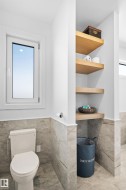 Bathroom featuring marble-look wall and floor tiling, a white toilet, a white framed window, and built-in wood-finish shelving - 