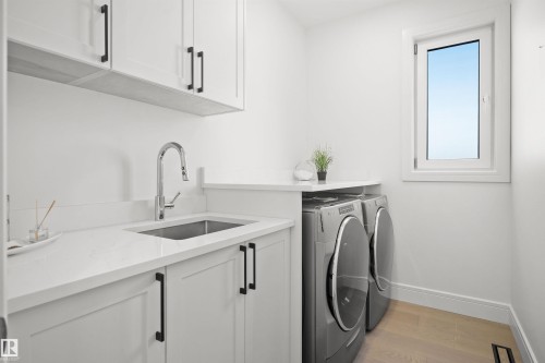 Dedicated laundry area featuring a stainless steel undermount sink, a chrome gooseneck faucet, light-toned wood-finish flooring, white cabinetry with black hardware, and a white framed window - 12304 62 Ave Nw, Edmonton, AB - Indoor Photo Showing Laundry Room