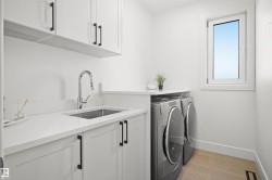 Dedicated laundry area featuring a stainless steel undermount sink, a chrome gooseneck faucet, light-toned wood-finish flooring, white cabinetry with black hardware, and a white framed window - 