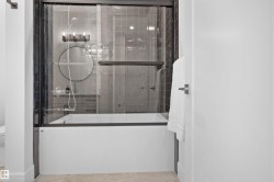 Bathtub with a glass enclosure, dark gray subway tile surround, and a coordinating dark gray vanity with a round mirror - 