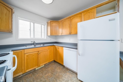 Kitchen featuring wood-finish cabinetry, a double basin stainless steel sink, white appliances, and a window above the sink - 12041/43 67 St Nw Nw, Edmonton, AB - Indoor Photo Showing Kitchen With Double Sink