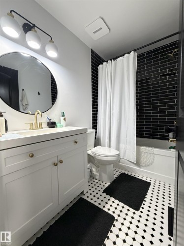 Bathroom featuring black subway tile shower surround, white vanity with gold-tone hardware, a circular mirror, and black and white hexagon tile flooring - 19F Meadowlark Village Nw, Edmonton, AB - Indoor Photo Showing Bathroom