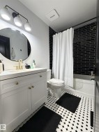 Bathroom featuring black subway tile shower surround, white vanity with gold-tone hardware, a circular mirror, and black and white hexagon tile flooring - 