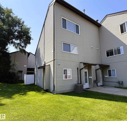 Rear exterior featuring light grey siding, multiple windows, a ground-level patio, and a grassy yard - 19F Meadowlark Village Nw, Edmonton, AB - Outdoor