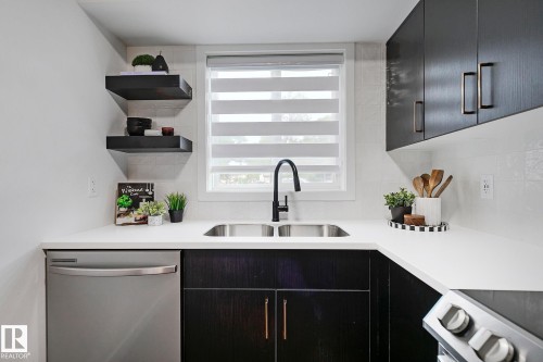 Kitchen featuring a double basin stainless steel sink with a matte black faucet, white countertops, dark wood-finish cabinetry with brass hardware, and a stainless steel dishwasher - 19F Meadowlark Village Nw, Edmonton, AB - Indoor Photo Showing Kitchen With Double Sink