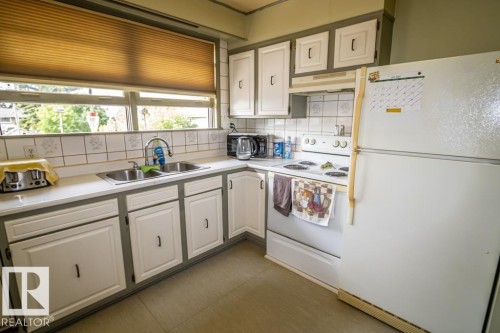 Kitchen featuring ample white cabinetry with dark hardware, a double basin stainless steel sink, tile backsplash, and a large window with privacy blinds - 5009 47 Ave, St. Paul Town, AB - Indoor Photo Showing Kitchen With Double Sink