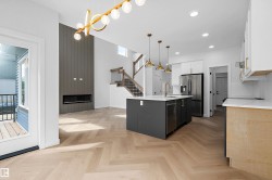 Open-concept living space showcasing herringbone wood-finish flooring, a central kitchen island with black cabinetry, stainless steel appliances, white quartz countertops, and pendant lighting - 