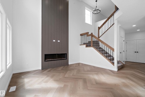 High ceilings with a prominent fireplace feature wall, light wood-finish herringbone flooring, and a staircase with wood handrails and black balusters - 1737 Crossbill Loop Nw, Edmonton, AB - Indoor Photo Showing Other Room With Fireplace