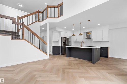 Open-concept living space with herringbone wood-finish flooring, a contemporary kitchen, a multi-level staircase with wood and iron railings, white upper cabinetry, and a dark-toned island with pendant lighting - 1737 Crossbill Loop Nw, Edmonton, AB - Indoor Photo Showing Kitchen With Upgraded Kitchen