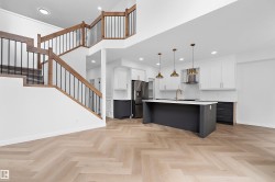 Open-concept living space with herringbone wood-finish flooring, a contemporary kitchen, a multi-level staircase with wood and iron railings, white upper cabinetry, and a dark-toned island with pendant lighting - 