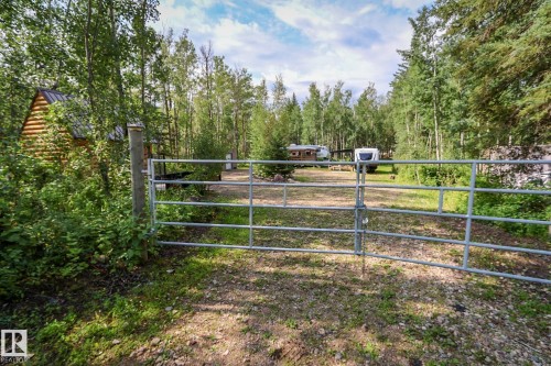 Gated entry to a property featuring a log cabin structure with a dark roof, surrounded by mature trees and natural landscaping - 200 12272 Township Road 602, Rural Smoky Lake County, AB 