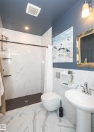 Modern bathroom featuring a wall-mounted toilet, pedestal sink, and a walk-in shower with white-veined tile walls and a dark shower pan - 