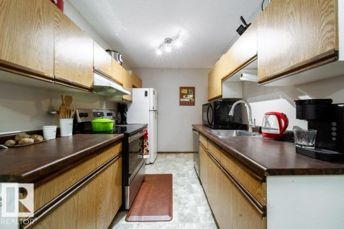 Galley kitchen featuring wood-finish cabinetry, dark countertops, white appliances, and a stainless steel sink with a chrome faucet - #307 6212 180 St Nw, Edmonton, AB - Indoor Photo Showing Kitchen