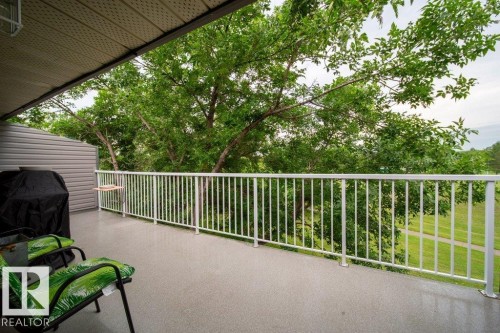 Expansive covered balcony featuring a textured gray surface, white metal railing, and mature green trees - #307 6212 180 St Nw, Edmonton, AB - Outdoor