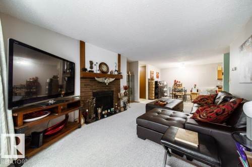 Spacious living area featuring a brick fireplace with a wood mantle, white textured ceilings, light-colored carpeting, and a recessed fireplace opening - #307 6212 180 St Nw, Edmonton, AB - Indoor Photo Showing Living Room With Fireplace