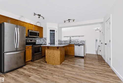 Kitchen featuring wood-finish flooring, a central island with a granite-style countertop, stainless steel appliances, a mosaic tile backsplash, and track lighting - #66 8304 11 Av Sw, Edmonton, AB - Indoor Photo Showing Kitchen With Stainless Steel Kitchen