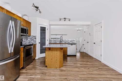 Kitchen featuring stainless steel appliances, wood cabinetry, wood-finish flooring, a kitchen island with a stone-finish countertop, and a tile backsplash - #66 8304 11 Av Sw, Edmonton, AB - Indoor Photo Showing Kitchen With Stainless Steel Kitchen
