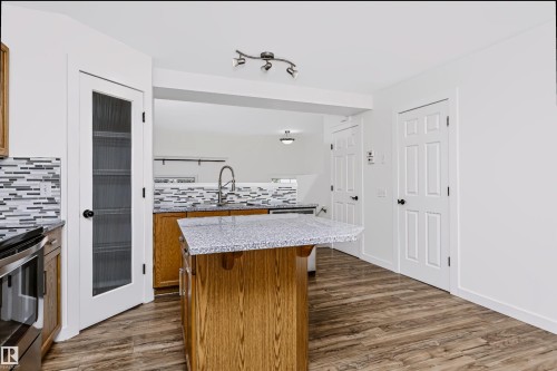 Kitchen featuring an island with a speckled countertop, wood-finish flooring, a mosaic tile backsplash, stainless steel appliances, and a glass-paneled pantry door - #66 8304 11 Av Sw, Edmonton, AB - Indoor Photo Showing Kitchen