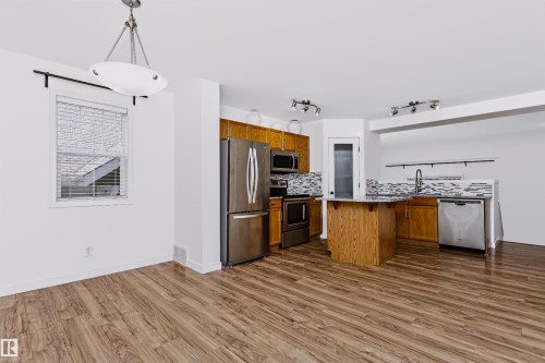 Open-concept kitchen and dining area featuring wood-finish flooring, stainless steel appliances, a center island with a granite-style countertop, full tile backsplash, and a contemporary pendant light fixture - #66 8304 11 Av Sw, Edmonton, AB - Indoor Photo Showing Kitchen With Stainless Steel Kitchen