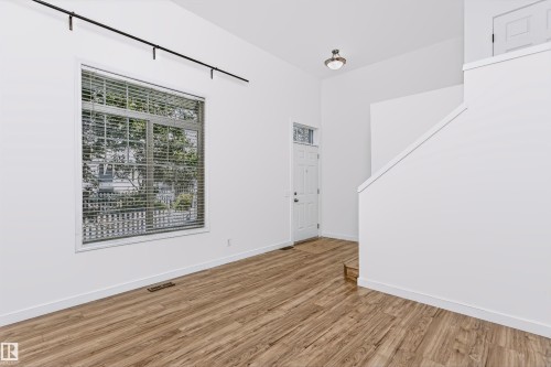 Open-concept living space featuring vaulted ceilings, wood-finish flooring, a contemporary window with blinds, a white entry door with a transom window, and a modern track lighting fixture - #66 8304 11 Av Sw, Edmonton, AB - Indoor Photo Showing Other Room