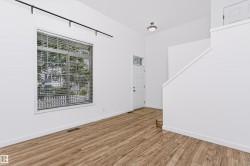Open-concept living space featuring vaulted ceilings, wood-finish flooring, a contemporary window with blinds, a white entry door with a transom window, and a modern track lighting fixture - 