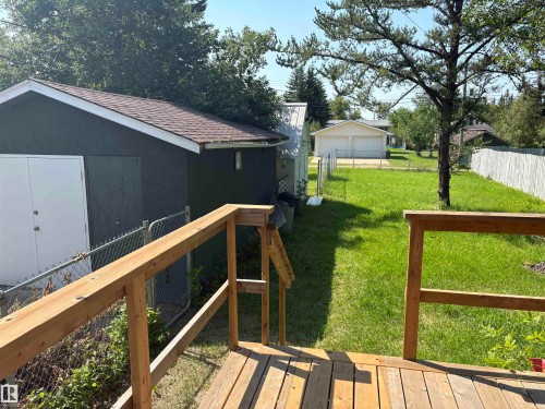 Rear deck with natural wood railing, overlooking a fenced yard featuring mature trees and a detached garage with dual doors - 4620 Maria Drive, Smoky Lake Town, AB - Outdoor