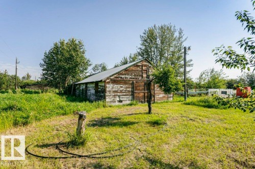 Wood-sided outbuilding with a metal roof, set within a grassy expanse featuring mature trees and additional structural elements - 55319 Rge Road 222, Rural Sturgeon County, AB 