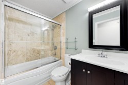 Bathroom featuring a white bathtub with sliding glass doors, natural stone-look tile surround, dark wood-finish vanity with an integrated sink, and a framed mirror with overhead lighting - 