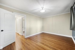 Spacious room featuring rich wood-finish flooring, crisp white baseboards and crown molding, a white ceiling light fixture, and a solid white interior door with a black handle - 
