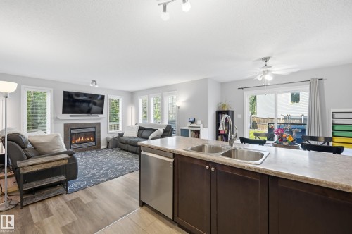 Open-concept living space with wood-finish flooring, a stainless steel dishwasher, an island with a double basin sink, a fireplace with a mantel, and sliding patio doors - 3606 Whitelaw Bay Nw, Edmonton, AB - Indoor With Fireplace