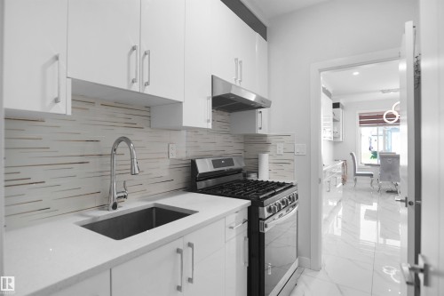 Modern kitchen featuring white cabinetry with silver hardware, a stainless steel sink with a gooseneck faucet, light-toned countertops, and a patterned tile backsplash - 4003 39 Street, Beaumont, AB - Indoor Photo Showing Kitchen With Upgraded Kitchen
