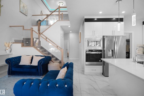 Open-concept living space featuring a modern staircase with glass panels and wood handrails, a contemporary kitchen island with a sink, white cabinetry, a built-in oven, and a stainless steel refrigerator - 4003 39 Street, Beaumont, AB - Indoor Photo Showing Kitchen With Upgraded Kitchen