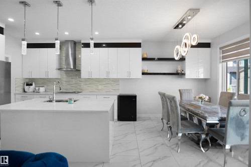 Modern kitchen featuring a large island with an undermount sink, pendant lighting, sleek white cabinetry, a stainless steel range hood, and marble-look tile flooring - 4003 39 Street, Beaumont, AB - Indoor
