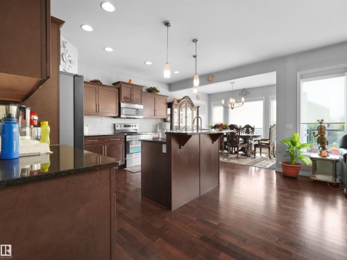 Open-concept kitchen featuring wood-finish flooring, dark wood cabinetry, a central island with dual pendant lighting, and stainless steel appliances - 1716 60 Street Sw, Edmonton, AB - Indoor Photo Showing Kitchen