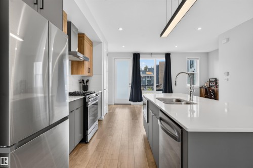Modern kitchen with stainless steel appliances, wood-finish flooring, white countertops, and contemporary lighting fixtures - #84 1304 Rutherford Rd Sw, Edmonton, AB - Indoor Photo Showing Kitchen With Stainless Steel Kitchen With Double Sink With Upgraded Kitchen