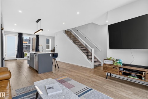 Open-concept living area featuring wood-finish flooring, a modern kitchen island with integrated appliances, a contemporary linear pendant light fixture, a staircase with metal balusters, and a wall-mounted display space - #84 1304 Rutherford Rd Sw, Edmonton, AB - Indoor