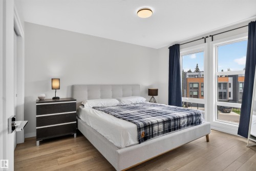 Bedroom featuring light wood-finish flooring, a contemporary flush-mount ceiling light, and two large windows with dark drapery - #84 1304 Rutherford Rd Sw, Edmonton, AB - Indoor Photo Showing Bedroom