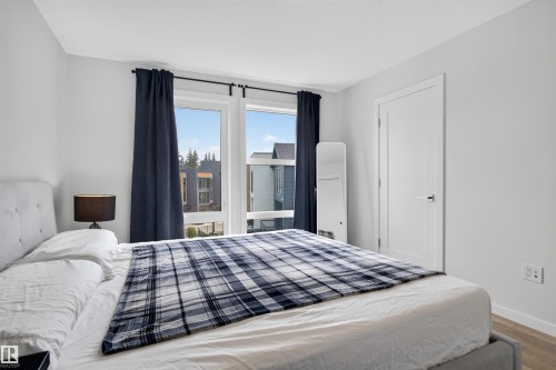 Bright room featuring large windows with dark curtains, light wood-finish flooring, a contemporary white door with a black handle, and crisp white walls - #84 1304 Rutherford Rd Sw, Edmonton, AB - Indoor Photo Showing Bedroom