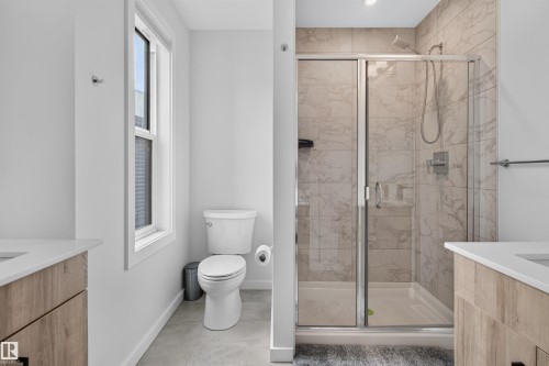Bathroom featuring a glass-enclosed shower with tile surround, a window providing natural light, a toilet, and dual wood-finish vanity cabinets with white countertops - #84 1304 Rutherford Rd Sw, Edmonton, AB - Indoor Photo Showing Bathroom