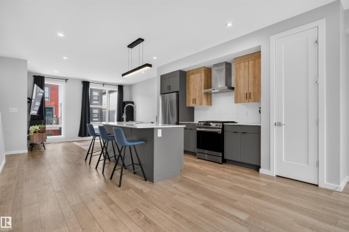 Open-concept living space featuring wood-finish flooring, a modern kitchen with a large island, stainless steel appliances, and sleek cabinetry - #84 1304 Rutherford Rd Sw, Edmonton, AB - Indoor Photo Showing Kitchen With Upgraded Kitchen