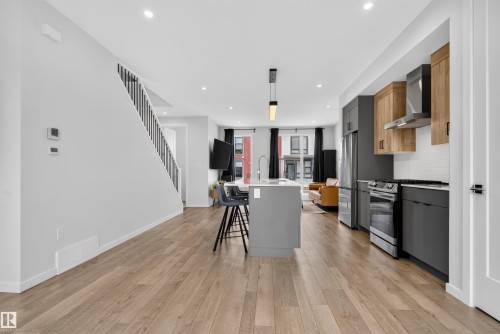 Open-concept interior featuring wood-finish flooring, a contemporary kitchen island with seating, stainless steel appliances, and a modern staircase with black railings - #84 1304 Rutherford Rd Sw, Edmonton, AB - Indoor Photo Showing Kitchen