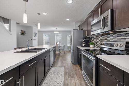 Kitchen featuring dark wood-finish cabinetry, stainless steel appliances, a double basin sink, light countertops, and a patterned tile backsplash - 12305 173A Avenue Nw, Edmonton, AB - Indoor Photo Showing Kitchen With Double Sink With Upgraded Kitchen