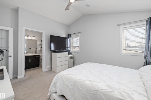 Carpeted room featuring a vaulted ceiling with a flush-mount ceiling fan, two windows with horizontal blinds, and a doorway to a bathroom with a dark wood vanity and light fixture - 12305 173A Avenue Nw, Edmonton, AB - Indoor Photo Showing Bedroom